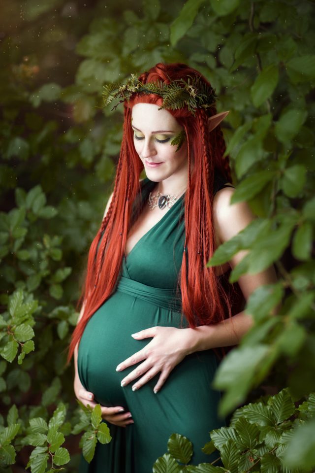 Babybauchshooting als Waldelfe im Zauberwald. Schwangere Frau mit roten langen Haaren und grünem Kleid. Fantasyfotografie.
